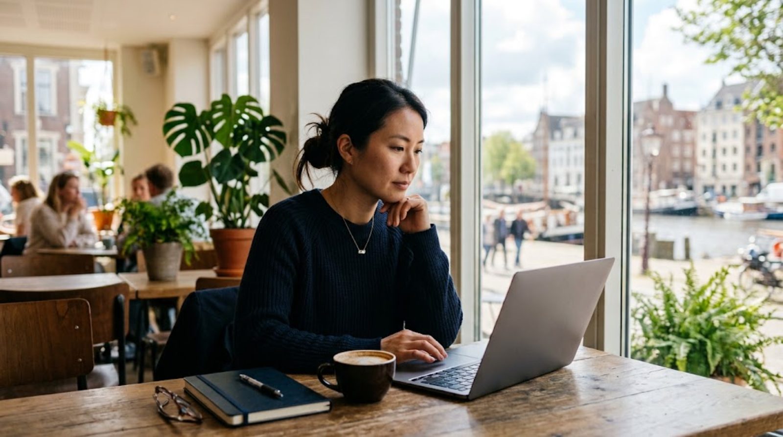 An international school teacher in their thirties working calmly on a laptop by a sunlit cafe window while traveling, rethinking income and how they work, a fully remote way of working unbound by location