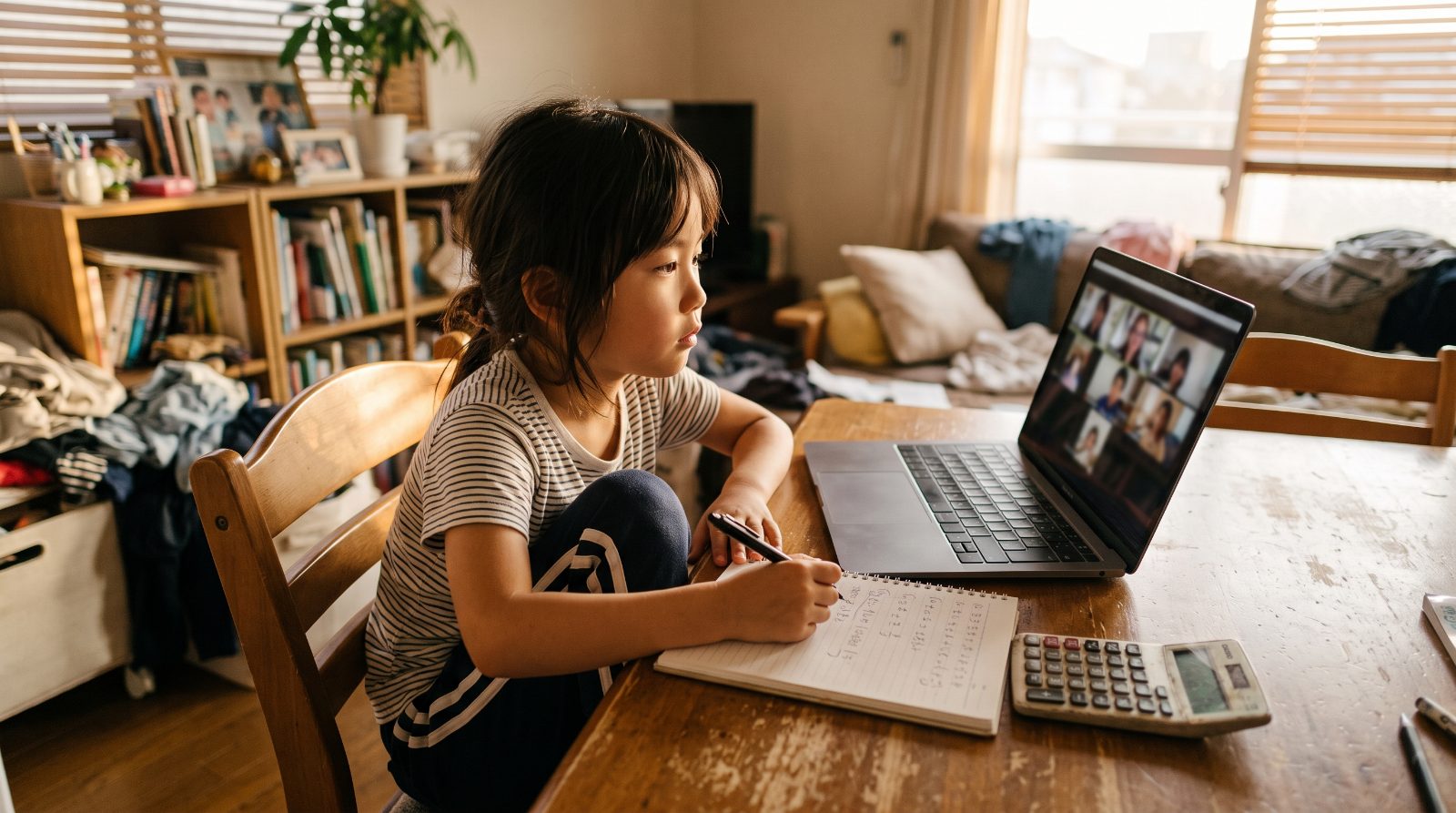 日本の家庭のリビングで子どもがノートパソコンでオンラインのインター授業を受け、机の上に6年間の費用を書き出したノートが置かれた温かい情景